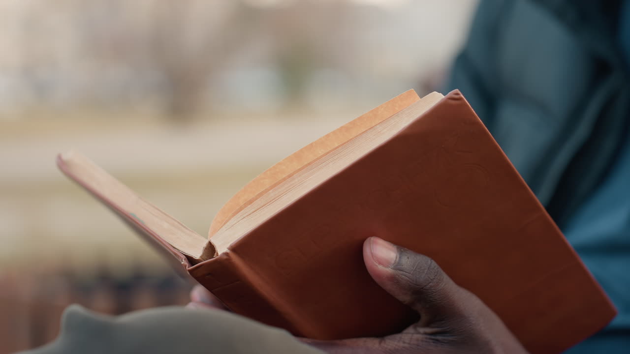 Close up of person reading hardcover book outdoors, holding cover with both hands while opening to new page, wearing warm jacket, seated in relaxed position with blurred park background