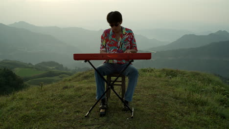 asian male musician in sunglasses plays piano at countryside mountain landscape wearing colorful vintage shirt