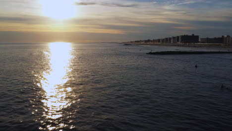 Cámara-De-Dron-De-Baja-Altitud-Tira-De-Una-Toma-Rodante-Con-Una-Puesta-De-Sol-Dorada-A-La-Vista---Surfistas-Abajo-Esperando-La-Ola-Perfecta