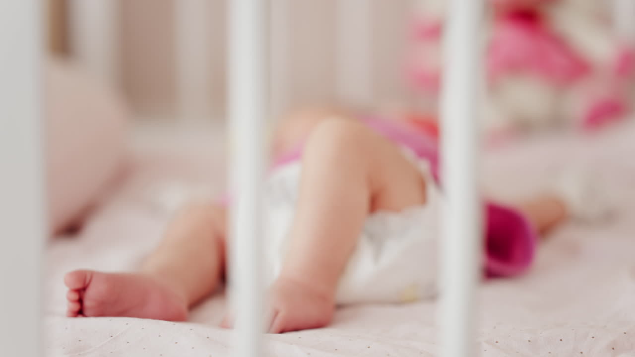Close up of a baby's legs and diaper as the infant rests in a crib, seen through the crib bars