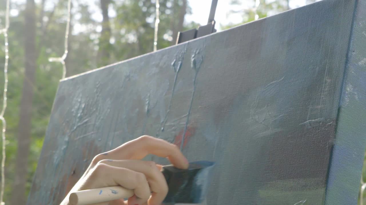 Female professional artist uses brush to paint abstract multicolored painting on canvas using brush strokes to the right, outside during the day, slow motion camera tilt up
