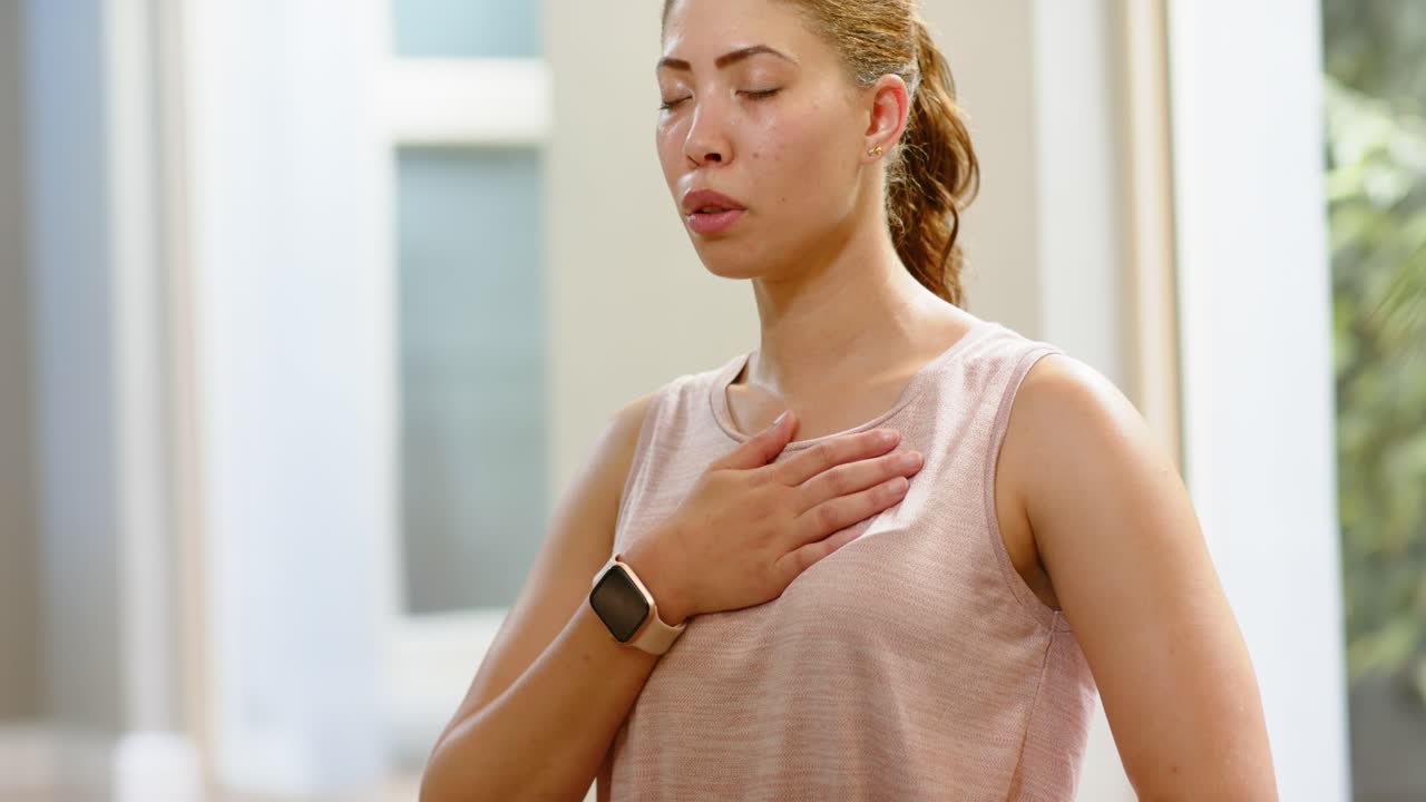 Donna che mette le mani sul petto e sull'addome, respirando per trovare calma a casa, indossando una canotta rosa e uno smartwatch.