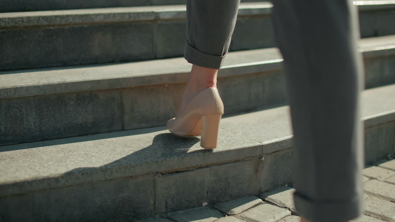 Lower leg view of woman walking in high heels on interlocked pavement ascending concrete steps, sunlight casting sharp shadow, focus on movement, footwear, business attire, and urban setting