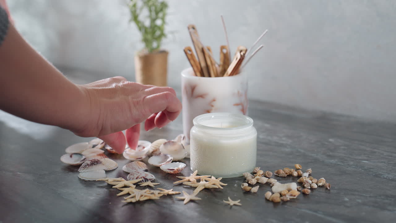 Hand view of woman arranging decorative materials including seashells, starfish, and pebbles on marble table near candle and plant pot in cozy indoor setting
