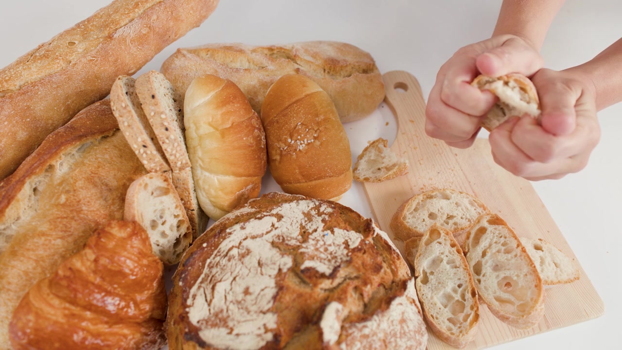 Close-up overhead shot of hands tearing crusty bread amidst baguettes, croissants, and rustic loaves.