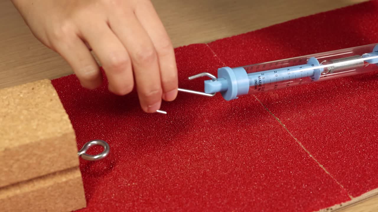 A hand conducts a friction test with a spring scale on a red textured surface, measuring force and movement