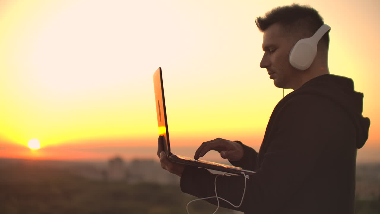 A man freelancer in headphones standing on the roof at sunset writes on the keyboard code pages. Little business. Listen to music and work at the computer enjoying the beautiful view from the roof.