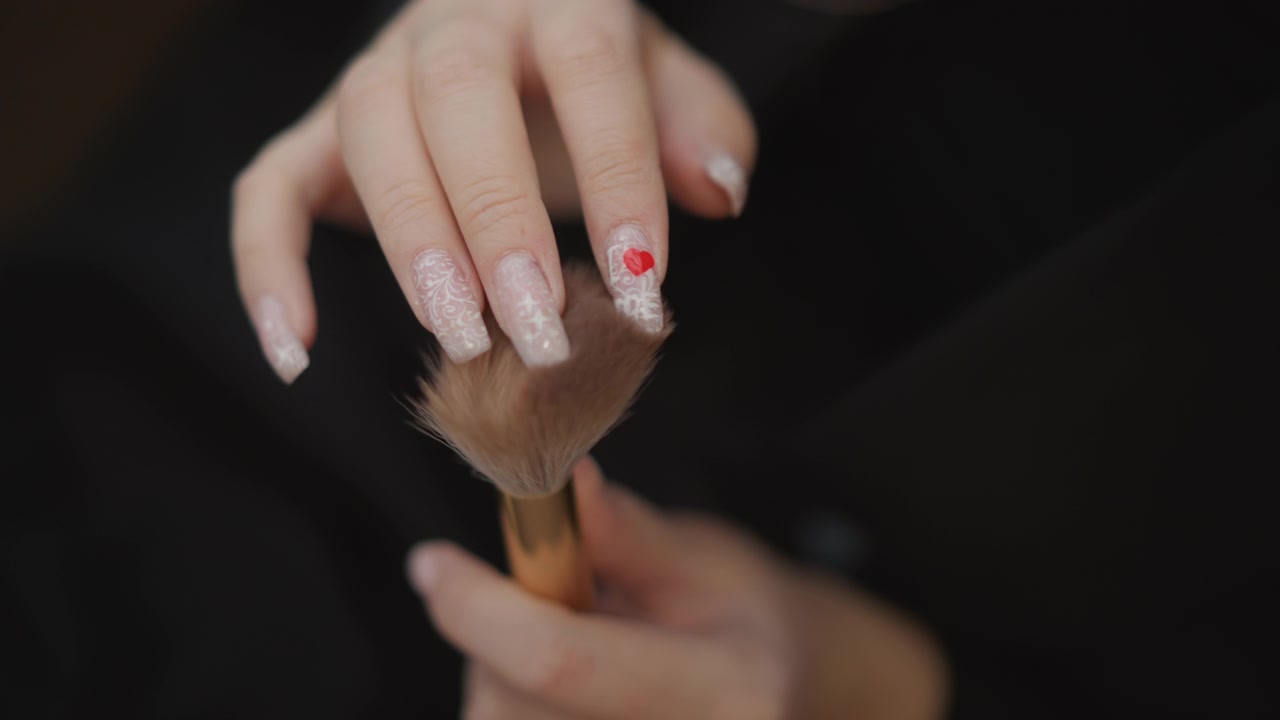 Close-up view shows manicured hands gently brushing powder across long square nails, with soft bristle motion, glossy glitter finish, red accents, and fine dust settling on skin and surface
