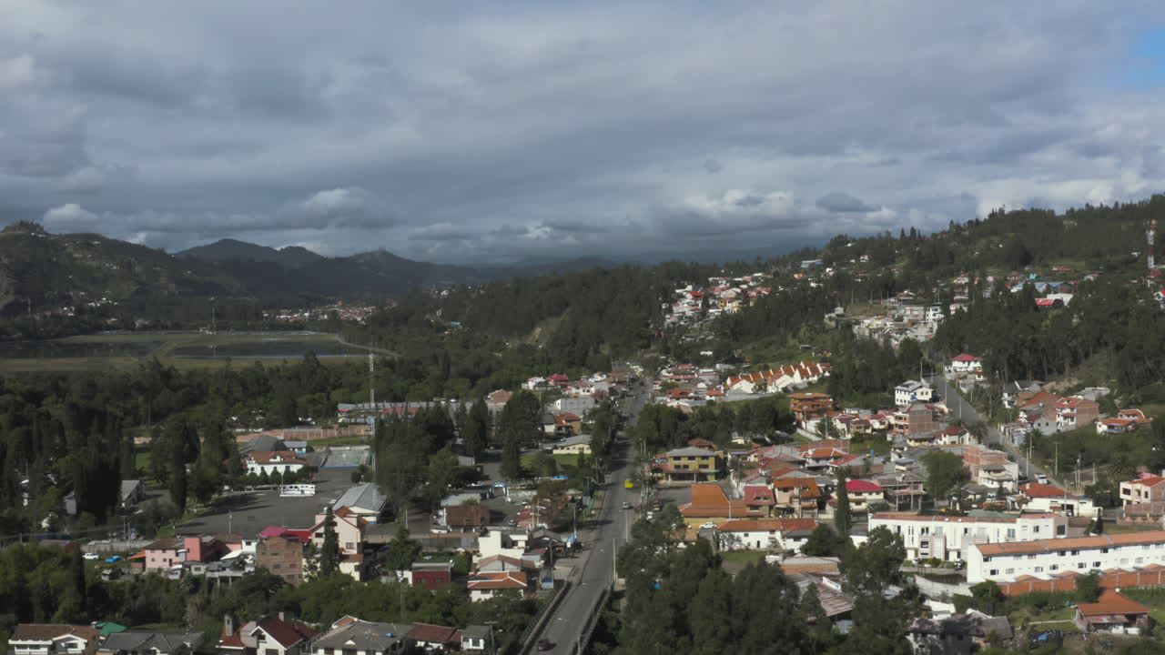 Aerial drone shot showing a residential area with houses scattered among green areas and mountains in the background.