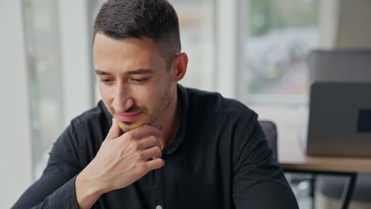 Pensive focused brunet man sitting at desk in front of laptop. Businessman thinking over something looking at computer screen. Close up. Blurred backdrop.