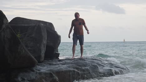 Man-Walking-Barefoot-On-The-Rock-With-Breaking-Waves-In-Dam-Trau-Beach,-Vietnam