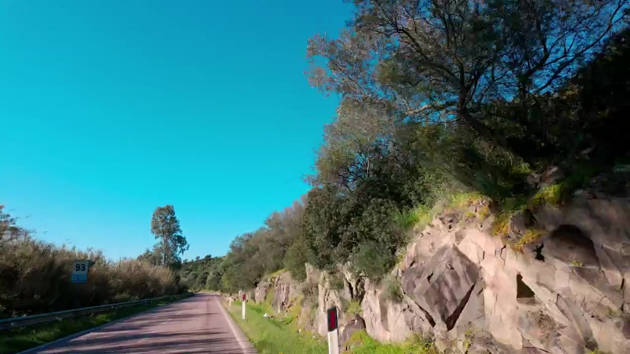 Una ripresa di guida in soggettiva lungo una strada tortuosa a Capo Carbonara, Sardegna, con alberi di un verde vibrante, scogliere rocciose e cieli azzurri limpidi.