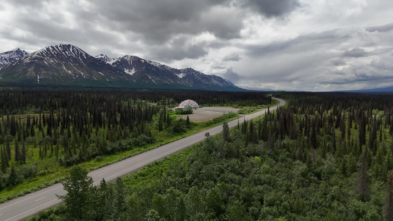 Drone Ascending over Parks Highway in Cantwell Alaska, Trees on sides of road, Talkeetna Mountains in Background with overcast sky and abandoned igloo shaped Motel in the distance