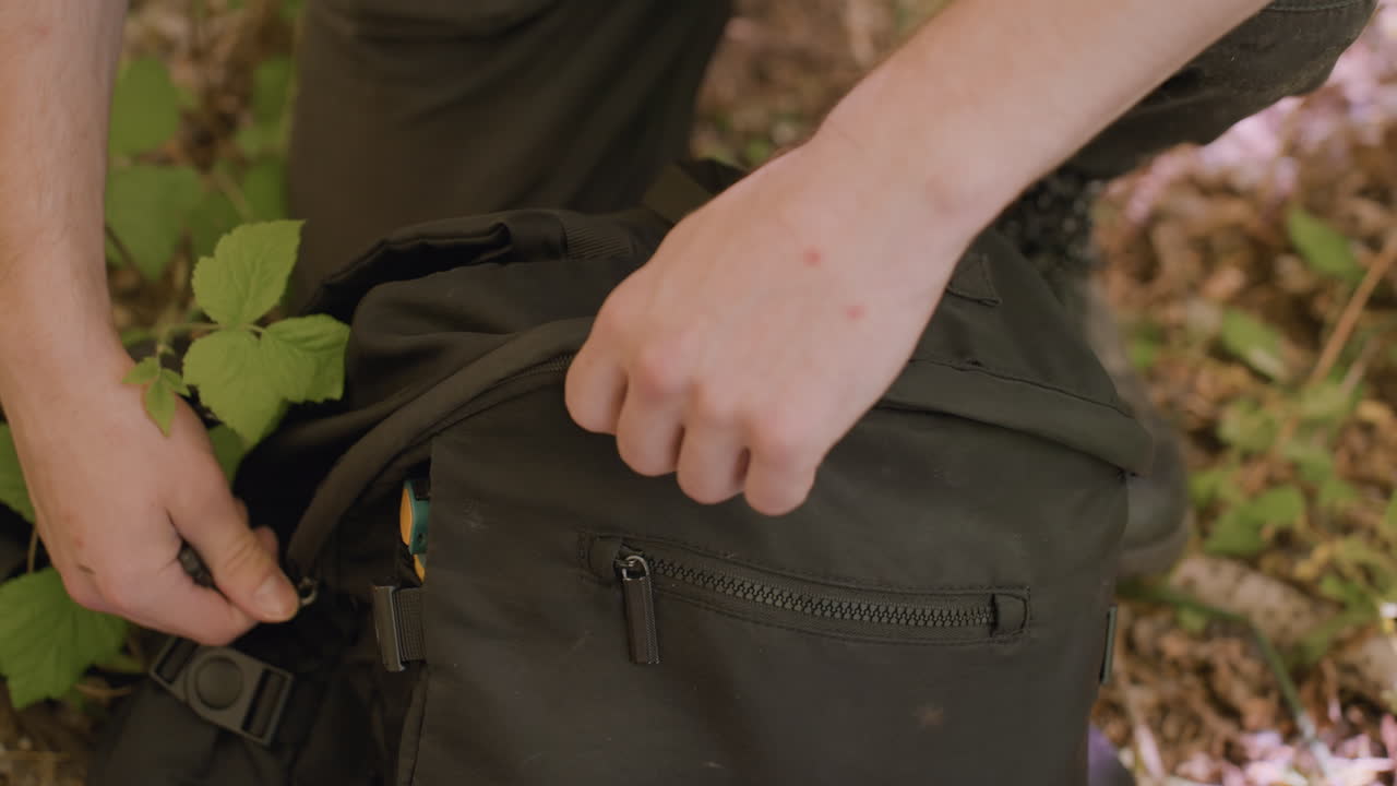 Tourist kneels in forest closing black bag, fingers pulling zipper while flies hover around gear and two fresh bite marks show on skin, amid fallen leaves and green undergrowth, ready to move on