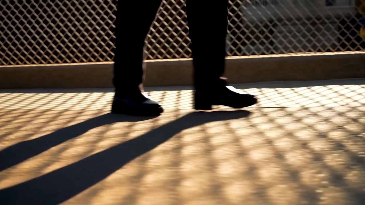 Close low-angle scene shows lower legs in polished dress shoes moving along concrete walkway beside chain-link fence, with long shadow patterns, warm sunlight, urban texture and quiet commute mood