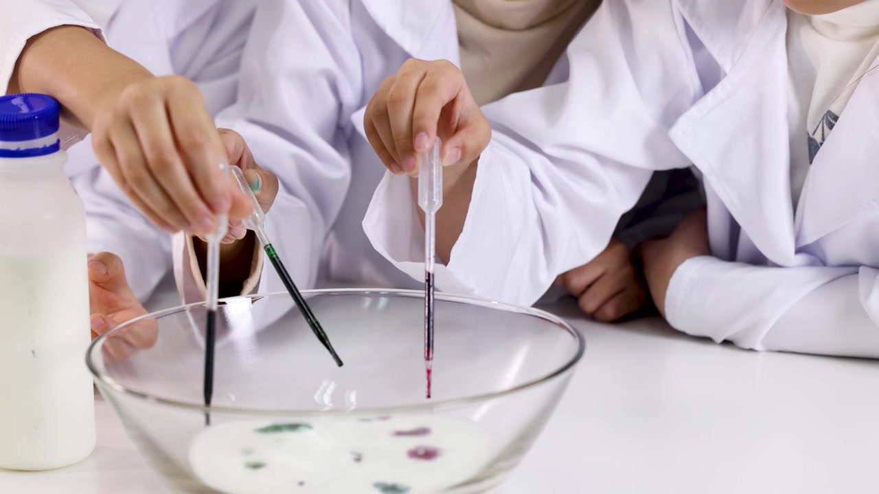 Close-up siswa menggunakan pipet untuk meneteskan pewarna makanan ke dalam susu selama percobaan sains di kelas