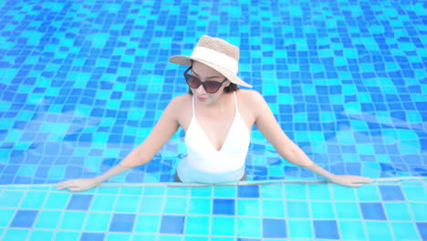 asian woman resting inside swimming pool, wearing white swimsuit, sunhat and sunglasses and leaning on pool's edge - high angle front view