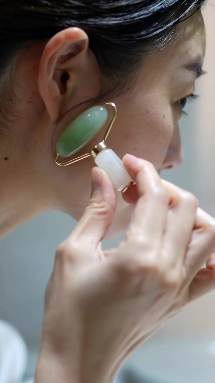 Vertical video: Leaning to mirror Asian woman rolling jade roller up cheek at vanity for skincare