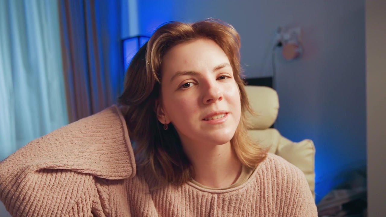 Medium view of young woman seated indoors in bedroom or living area, gently adjusting hair, touching neck, resting hand behind head, and keeping calm direct gaze in soft mixed light