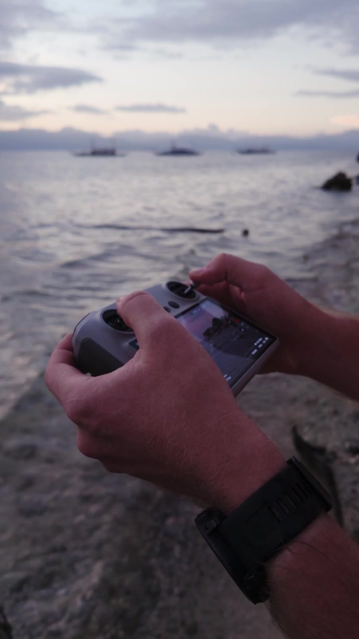 Inquadratura verticale delle mani di un uomo che tengono un controller per drone, con l'oceano sfocato sullo sfondo.