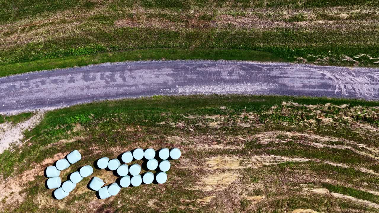 A top-down aerial drone shot flies over a harvested agricultural field in Latvia, showing numerous round hay bales wrapped in plastic for silage