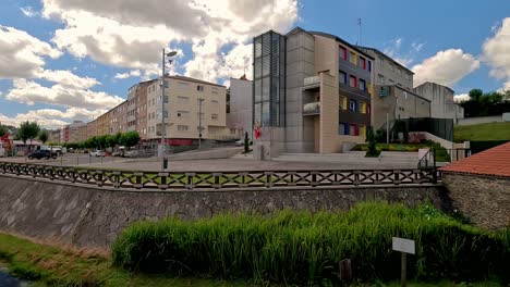 Salida-Del-Río-Subterráneo-Que-Cruza-El-Pueblo-Con-Edificios-Residenciales-De-Tamaño-Mediano-Y-El-Parque-Infantil-En-El-Centro-Un-Día-Soleado-Con-Nubes,-Tiro-Girando-A-La-Izquierda,-Ordes,-Galicia,-España