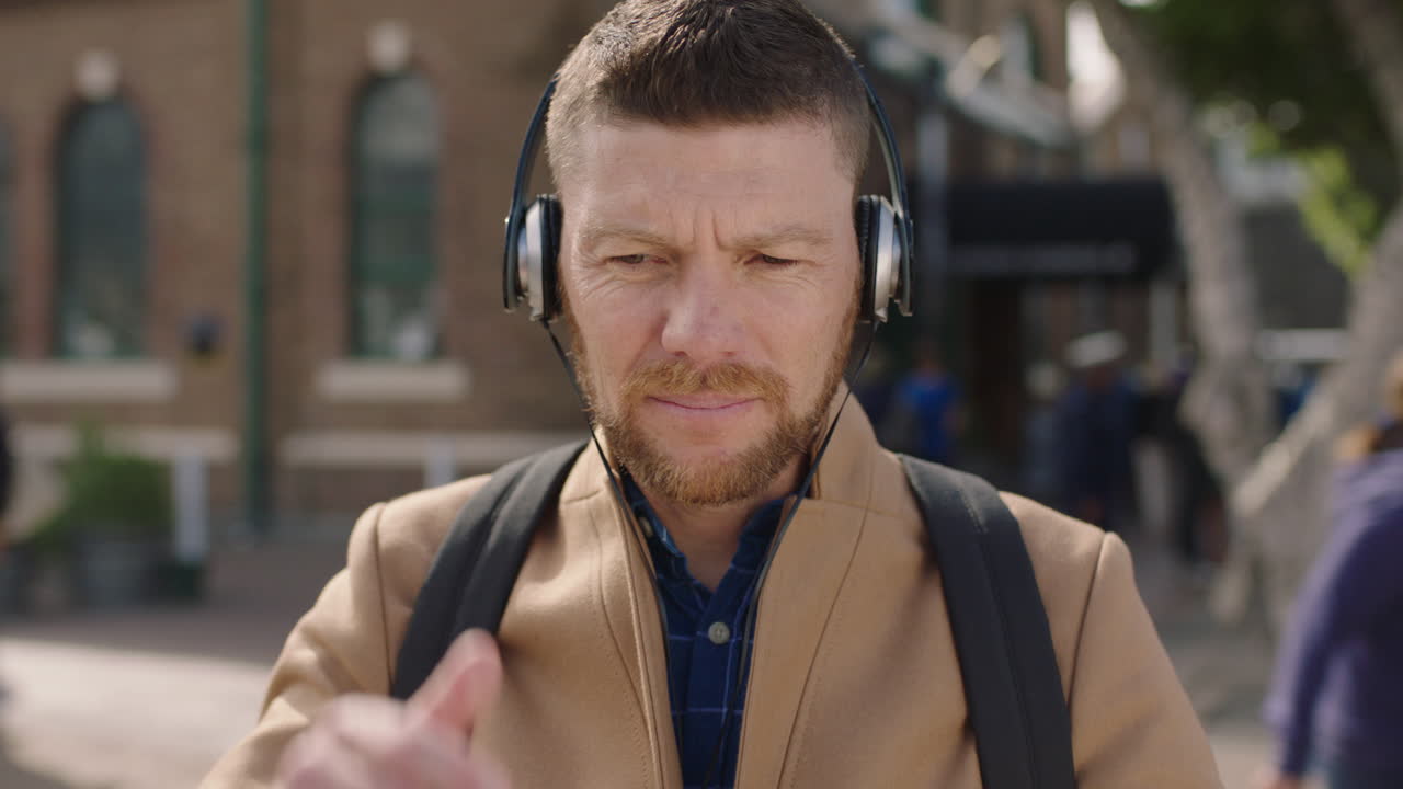 Nærbillede af charmerende kaukasisk mand, iført hovedtelefoner, der lytter til musik og tekster på sin smartphone i byen.