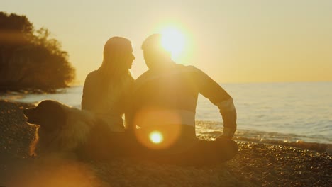 young couple with dog sitting on the lake, admiring the sunset