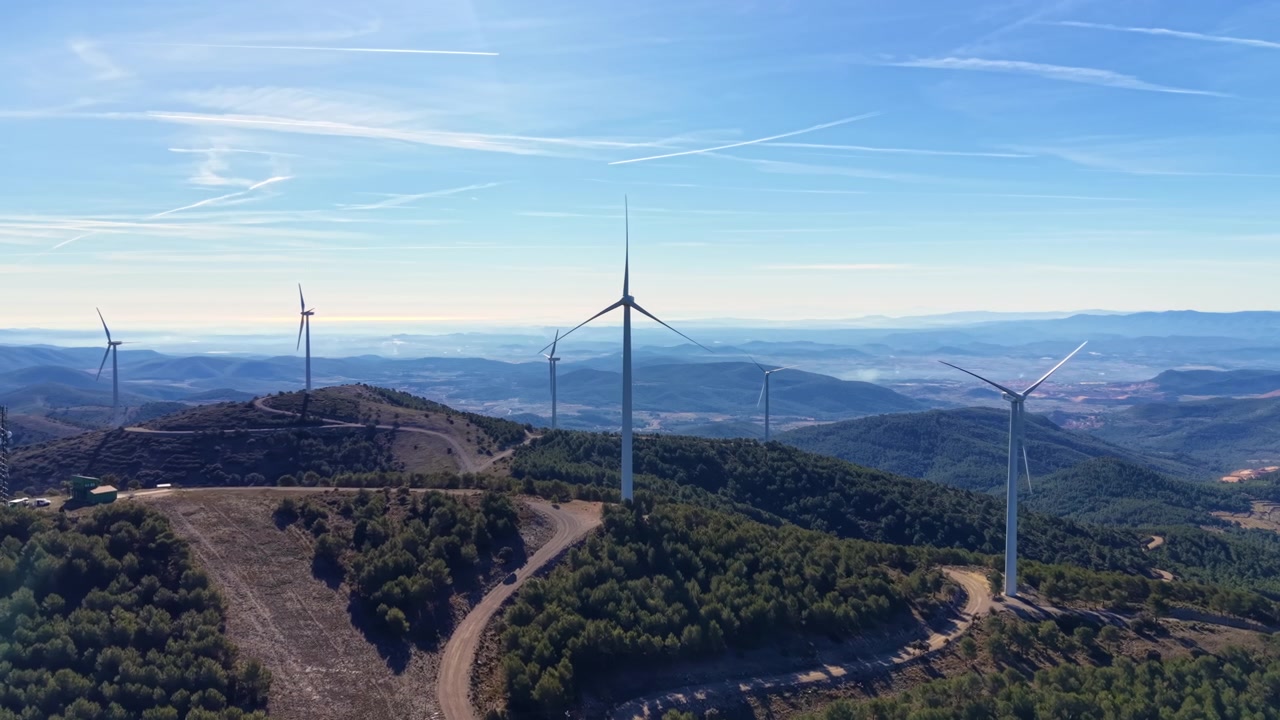 Vindmøller står stille på spanske bjergkamme under en klar blå himmel, omgivet af skovklædte bakker og et stort dallandskab, der strækker sig mod horisonten.