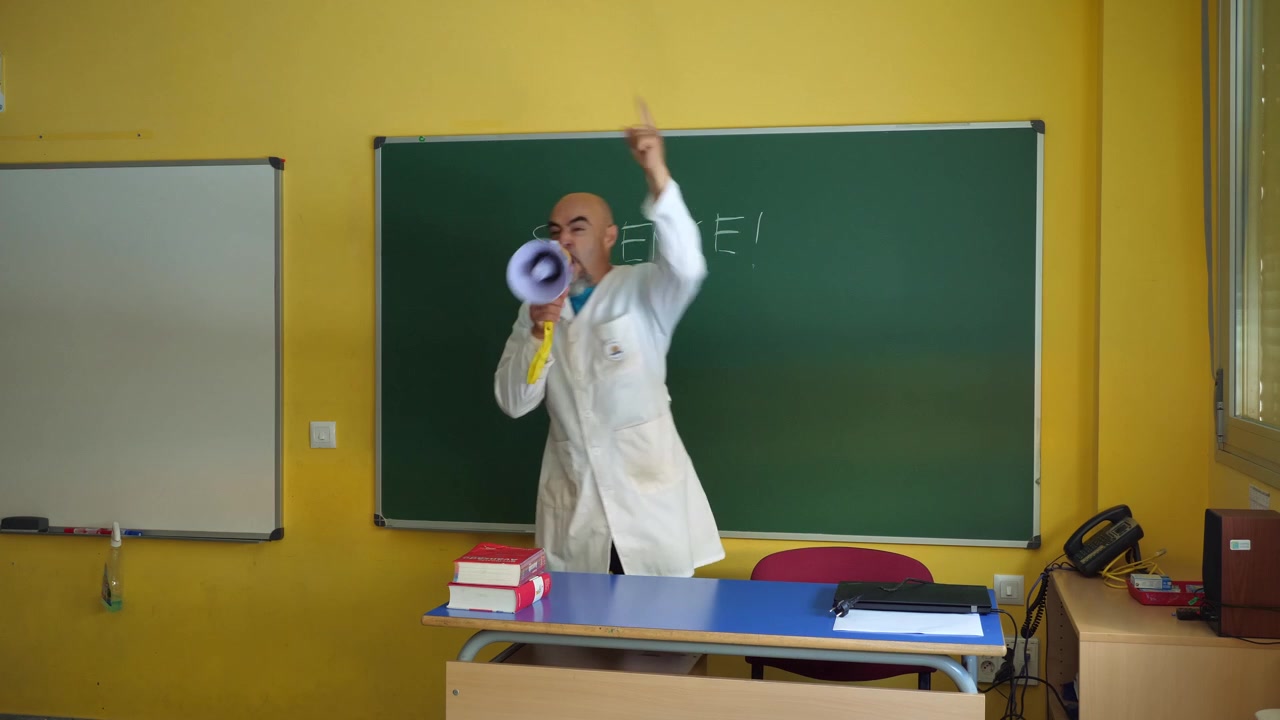 Inquadratura media di un insegnante maschio in camice bianco che scrive 'SILENZIO!' con il gesso su una lavagna verde. Si gira con un'espressione seria, afferra un megafono giallo dalla sua scrivania e urla.