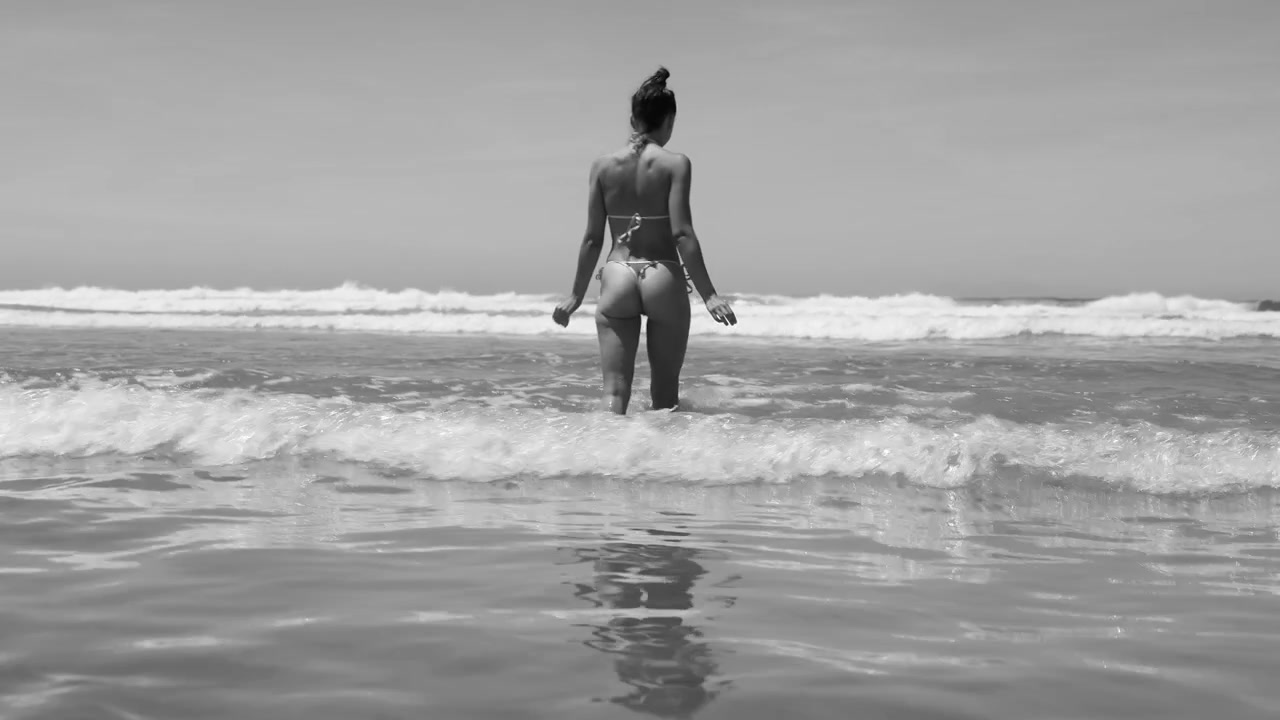 Kvinde i g-strengsbikini går ud i havet i sort-hvid
