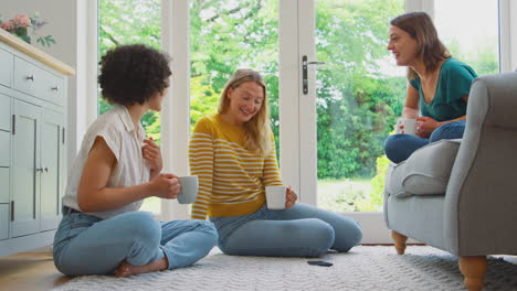 group of female friends relaxing in lounge at home looking at mobile phone and drinking coffee