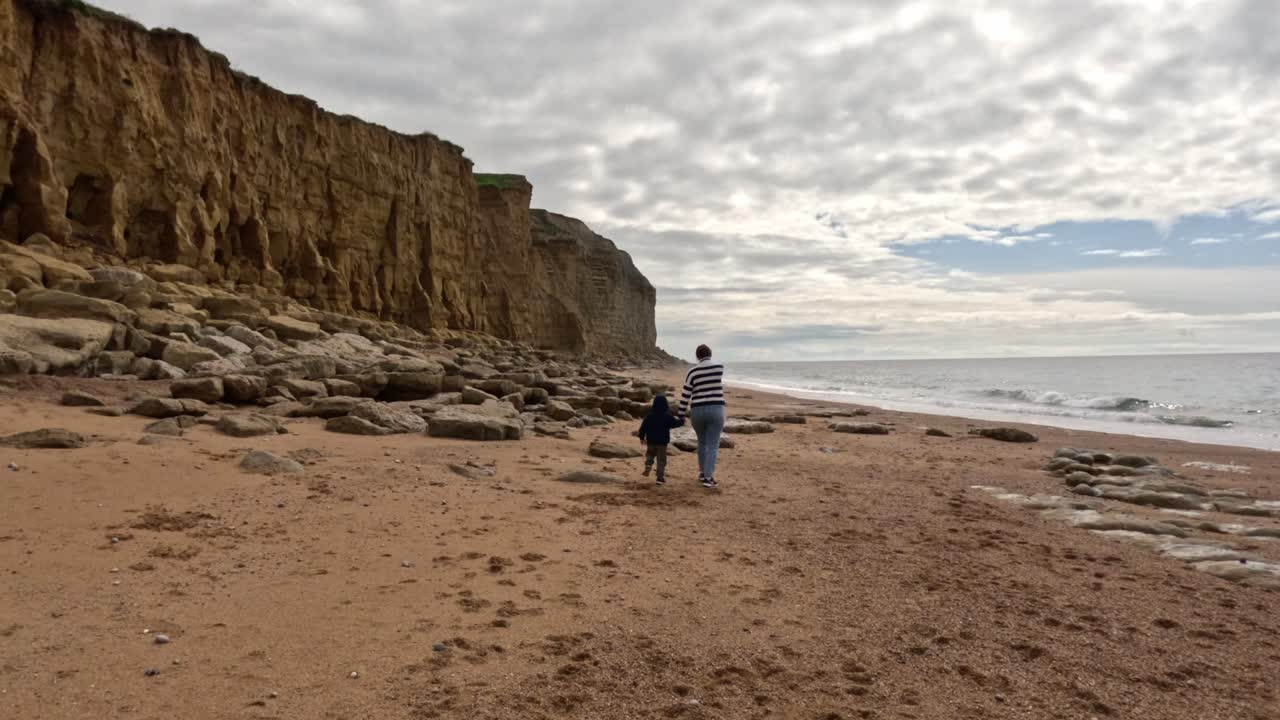 Captured in slow motion a mother and young, son walking on a Jurassic Coast beach, Dorset. Distinctive cliffs on the south coast of England on the left of the shot with rocks at the bottom, sea right.