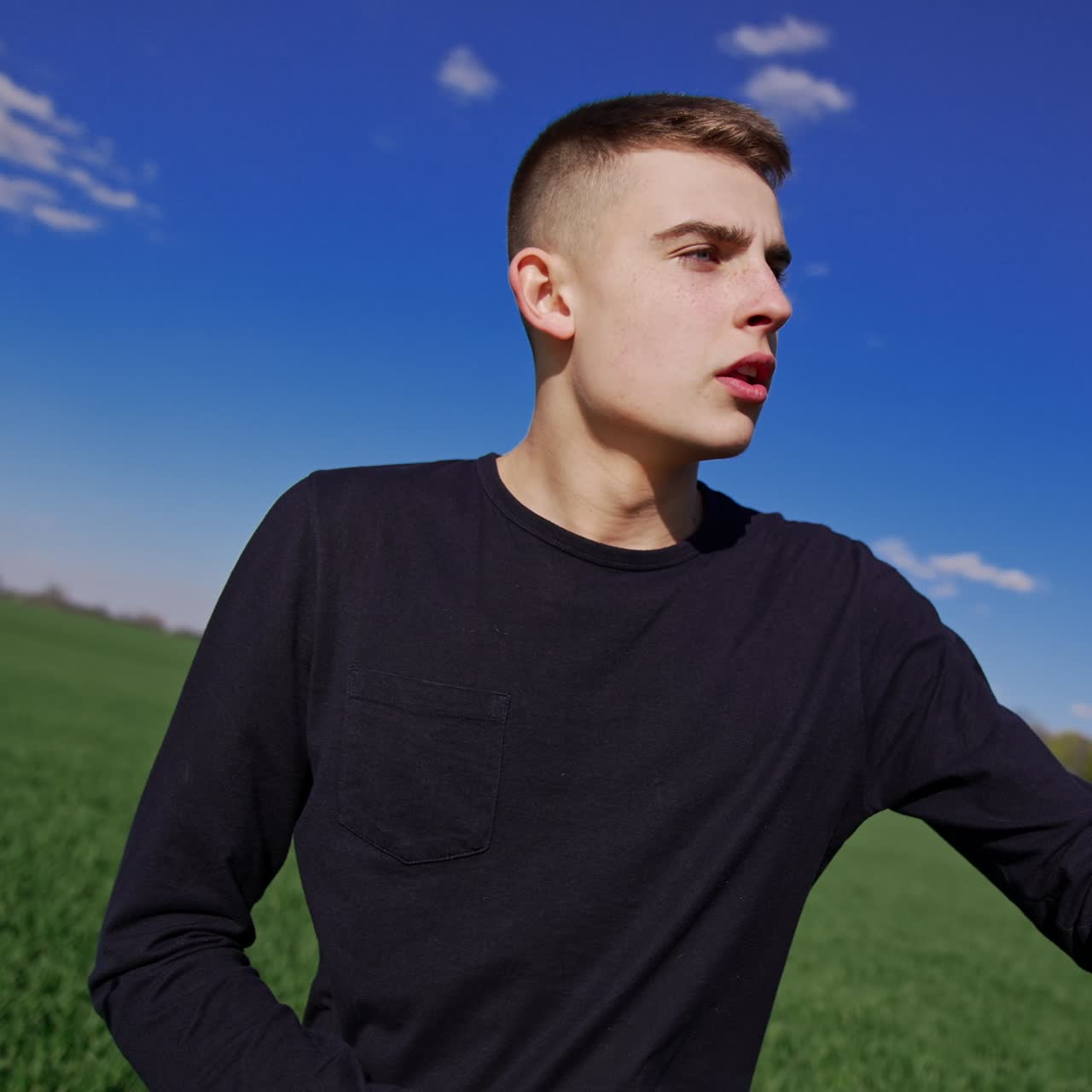 Young guy playing drums turns the drumsticks in his hand. Musician sitting outdoors in the green field under blue skies. Low angle view