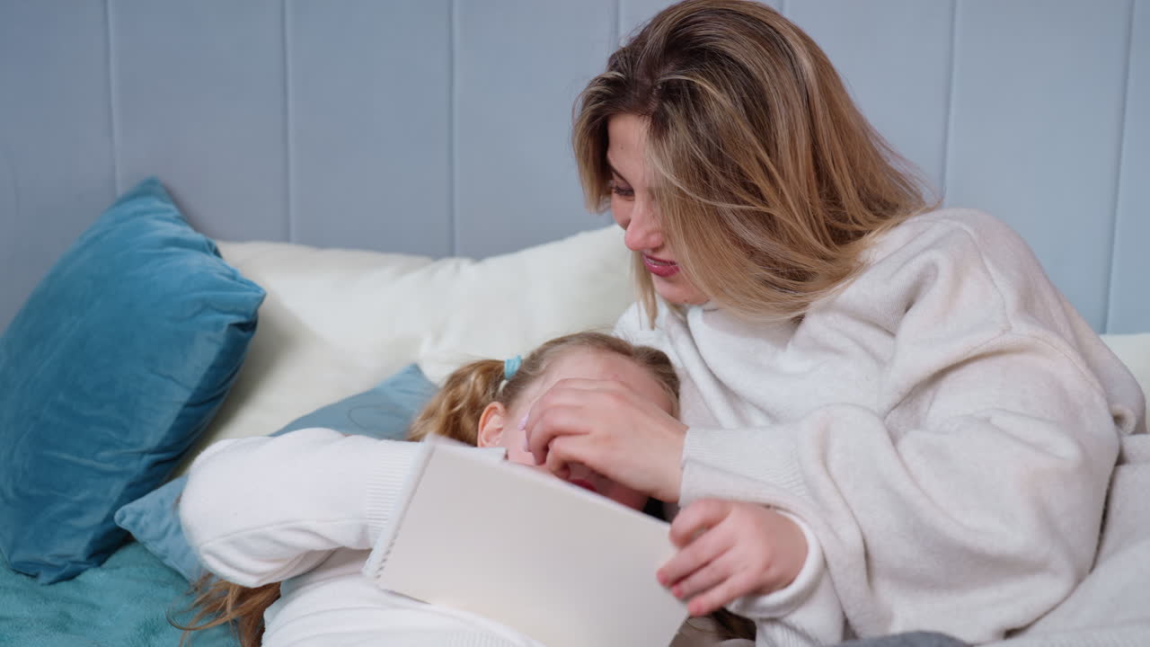 Smiling woman relaxing on bed in cozy sweater talking with cheerful girl, enjoying warm family bonding moment with pillows and blanket creating intimate atmosphere of happiness, playful connection
