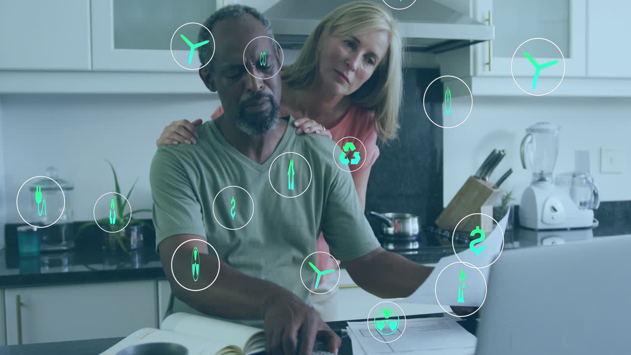 Animation of green energy concept icons against biracial couple calculating finances at home