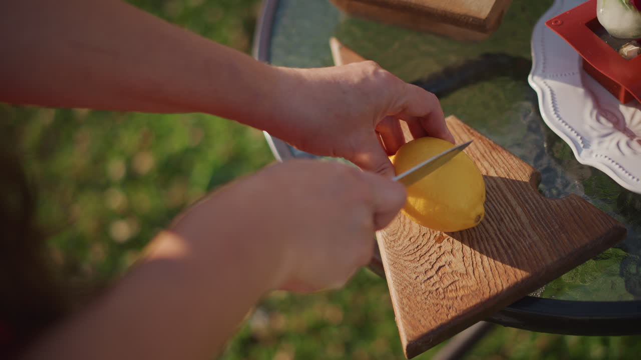 Limón en primer plano sobre tabla de cortar, manos blancas haciendo cortes precisos para guarnición de cóctel en patio soleado, vaso frío y hielo cerca, preparación de bebida veraniega vibrante con detalles brillantes de mixología