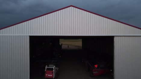 Gran-Hangar-Blanco-Con-Maquinaria-Agrícola-En-El-Interior-Y-Una-Persona-De-Pie-En-El-Otro-Extremo-Durante-El-Anochecer