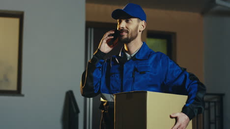 Atractivo-Cartero-Con-Traje-Azul-Y-Gorra-Con-Cartón-Percel-Hablando-Por-Teléfono-Móvil