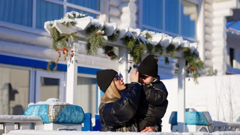 long-haired blonde woman wearing black jacket holds a baby boy in black jacket. mom and son wearing sunglasses on a sunny winter day. mother points at something ahead.