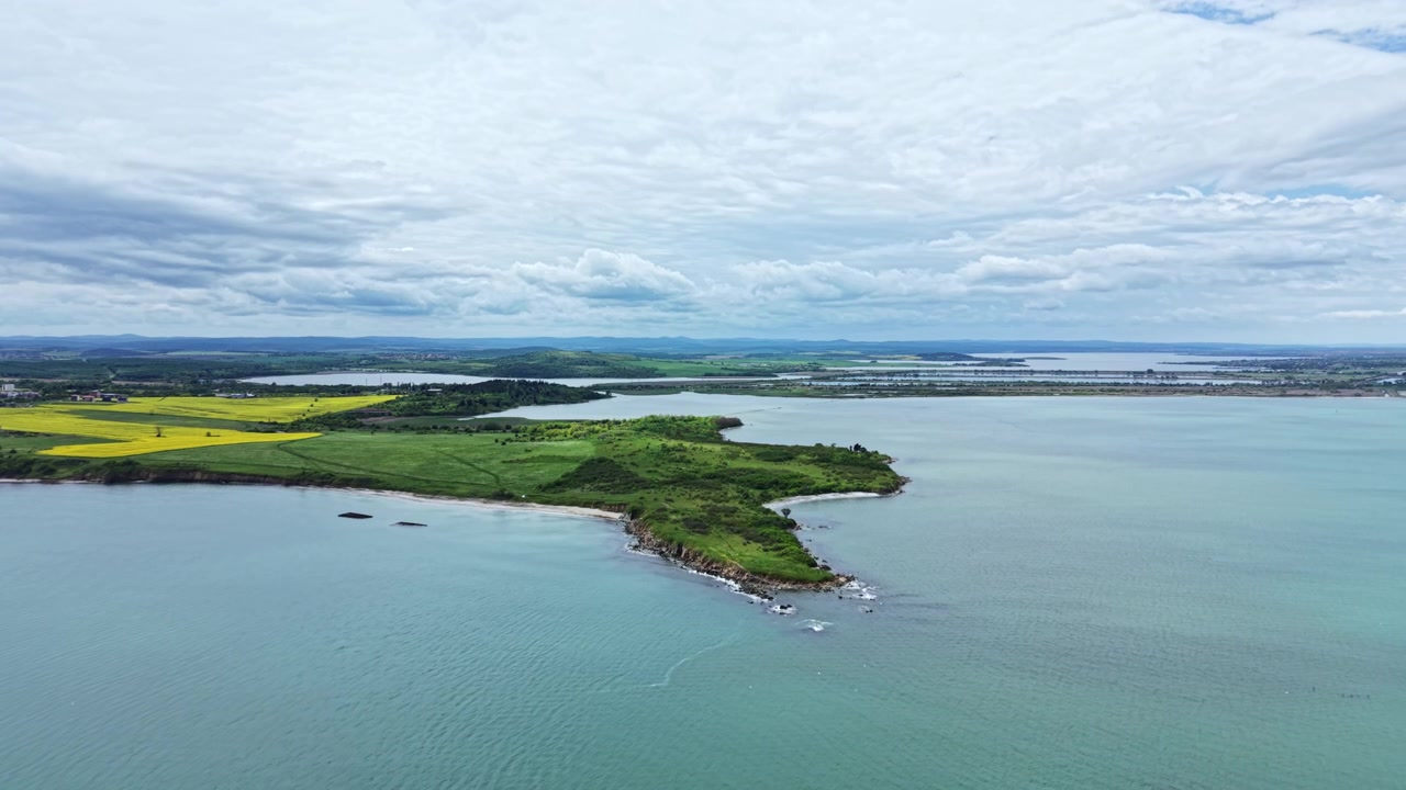 Naturskøn udsigt over et landskab, der viser vand, land og skyer på en overskyet dag med grønne områder og marker i det fjerne.