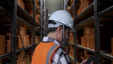 vista cercana de atrás de un ingeniero masculino asiático con casco de seguridad hablando en teléfono inteligente mientras está de pie en el almacén con estantes llenos de productos de entrega