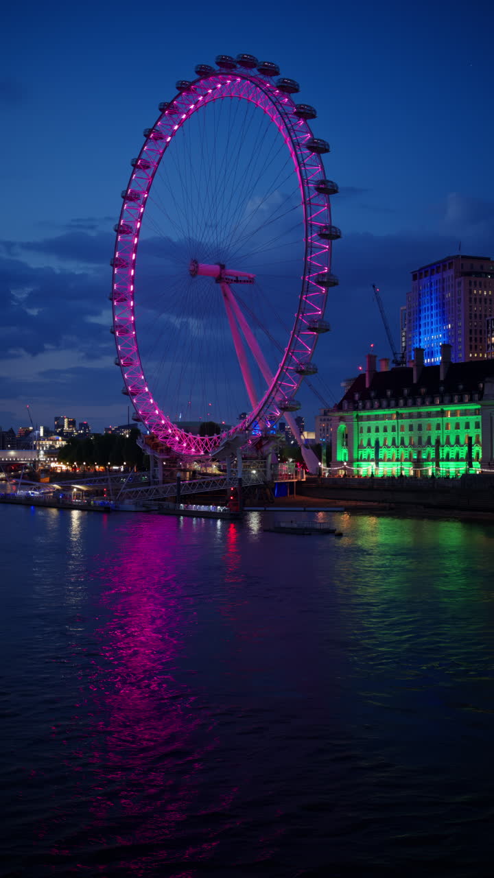 View of London Eye glowing pink and County Hall lit in green in the evening. Vertical, London