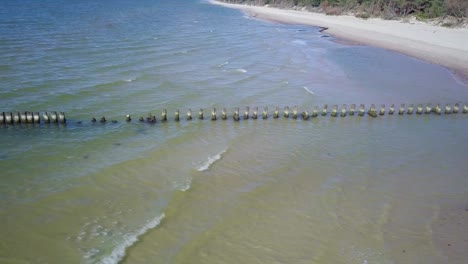 Aerial-birdseye-view-of-Baltic-sea-coast-on-a-sunny-day,-old-wooden-pier,-white-sand-coastline-damaged-by-waves,-coastal-erosion,-climate-changes,-wide-angle-drone-dolly-shot-moving-left