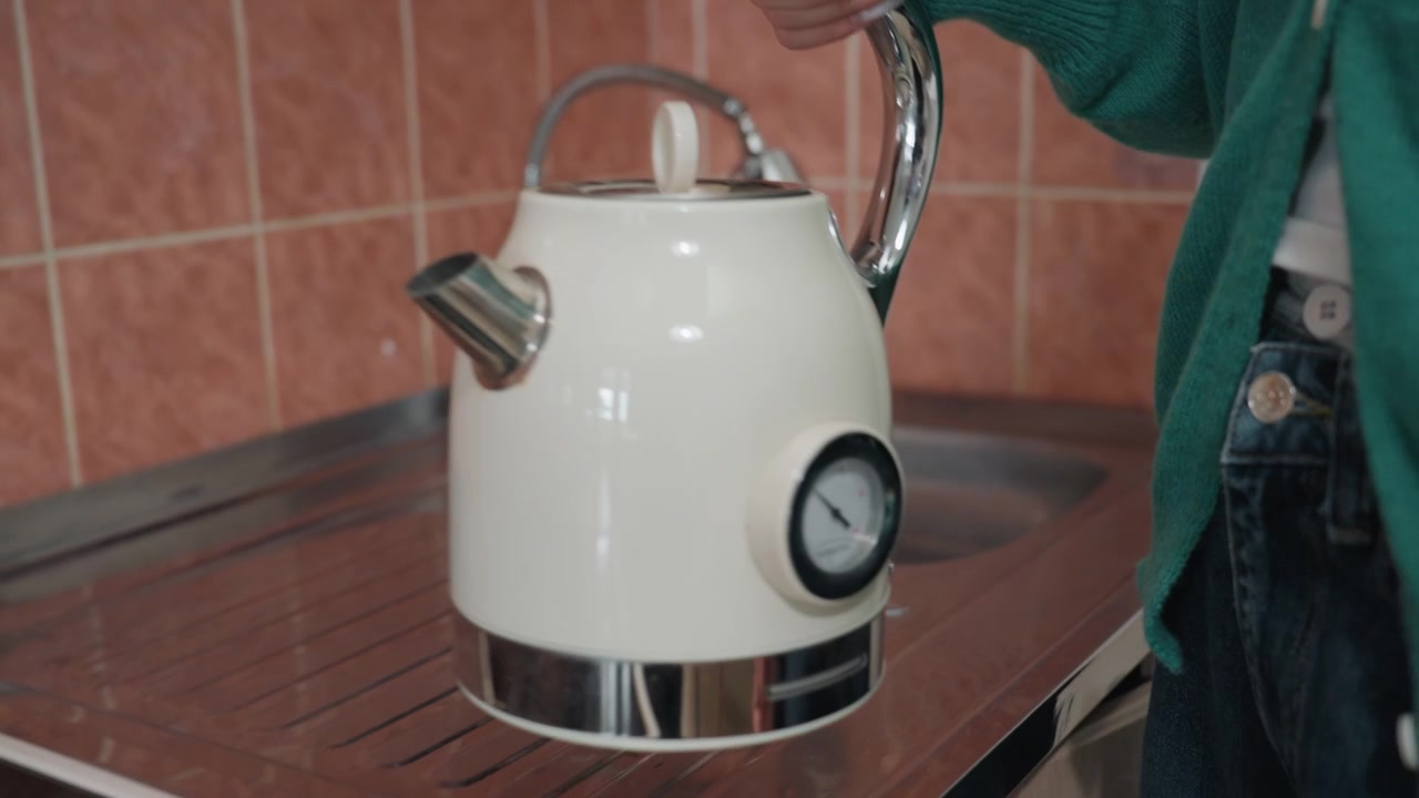 Proprietario di casa che posiziona un bollitore sul fornello nella cucina della nuova casa, le mani abbassano il coperchio e regolano la manopola. Bollitore bianco retrò con indicatore su piano cottura in ceramica, parete piastrellata e lavello con scolapiatti sullo sfondo, mentre disimballa.