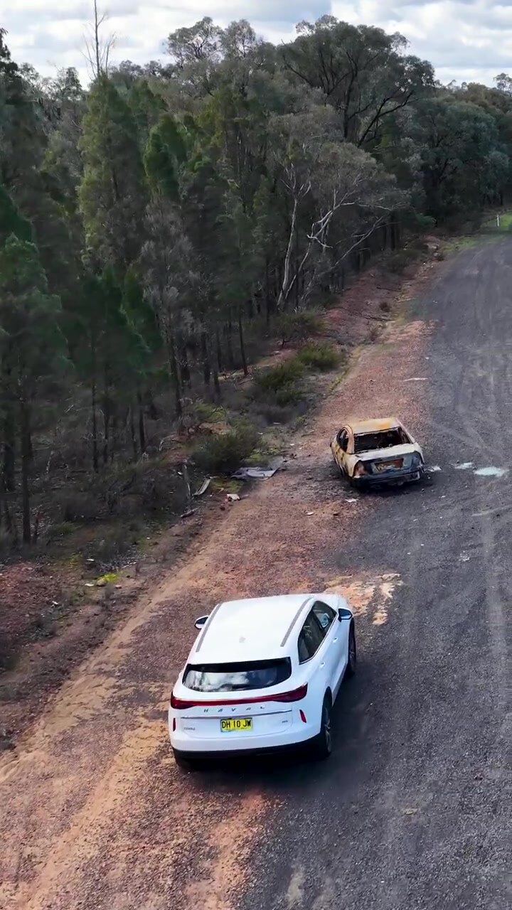 Vista aérea de un vehículo blanco pasando los restos de un coche abandonado y carbonizado en el Bosque Goonoo.