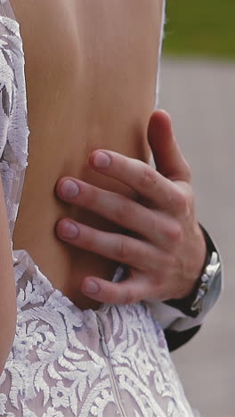 cu, slow motion: groom with wrist watch hugs gently bride in elegant white lacy wedding dress with bare back holding bouquet on street extreme closeup