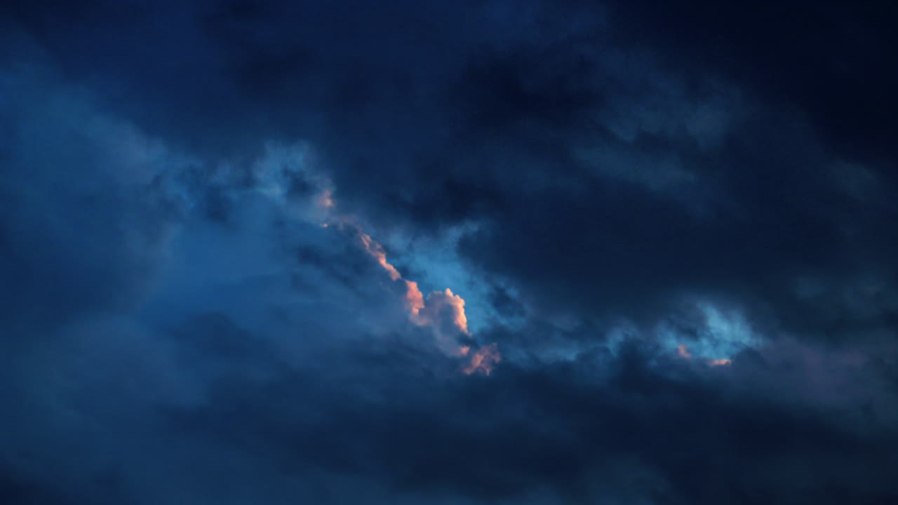 Moody dark blue clouds move across the sky at dusk, revealing a streak of warm light through the center