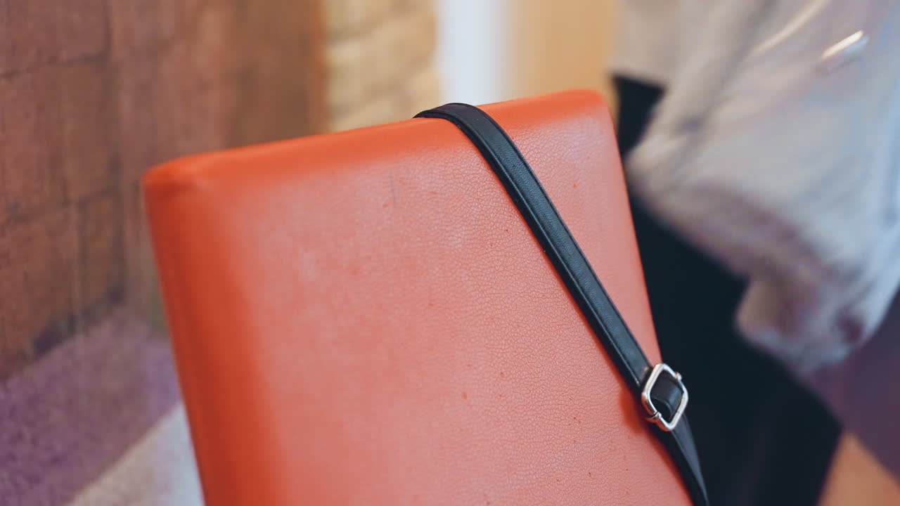 Borsa arancione all'interno del cappotto, mani in primo piano che infilano la borsa di pelle in una giacca calda, pendolare che regola la cinghia e la fibbia mentre entra in un corridoio urbano, superficie in pelle testurizzata, tonalità tenui, inverno accogliente