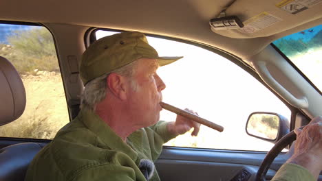 old man wearing cap drives in car, holding long cigar while talking animatedly